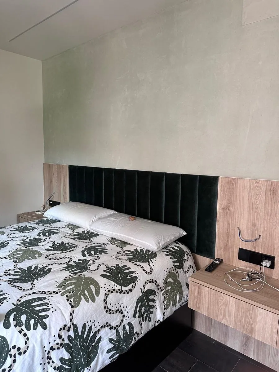 Dormitorio con una pared revestida en Estukum de tono verde suave, cabecero tapizado verde oscuro y cama con colcha de hojas
